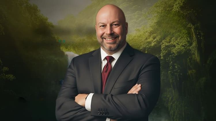 Portrait of a smiling bald man in a dark suit and red tie standing before a lush forest waterfall backdrop.