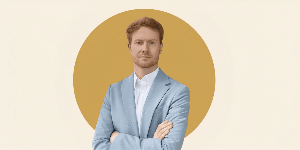 Man in a light blue suit standing with arms crossed against a minimalist gold circular background.