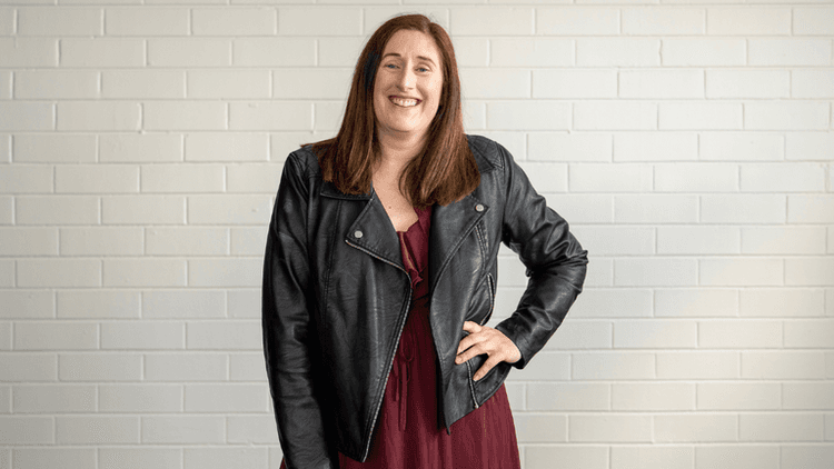 Woman smiling in a leather jacket and burgundy dress against a white brick wall