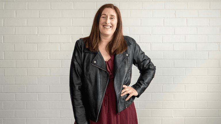 Woman smiling in a leather jacket and burgundy dress against a white brick wall