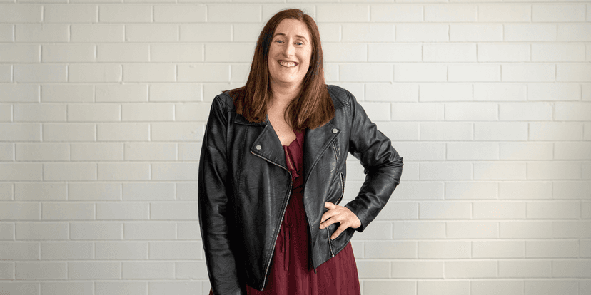 Woman smiling in a leather jacket and burgundy dress against a white brick wall