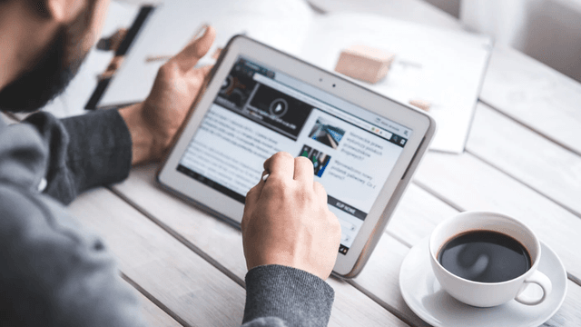 Person using a tablet to read news while sitting at a table with coffee
