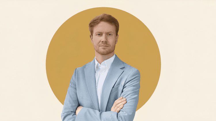 Man in a light blue suit standing with arms crossed against a minimalist gold circular background.