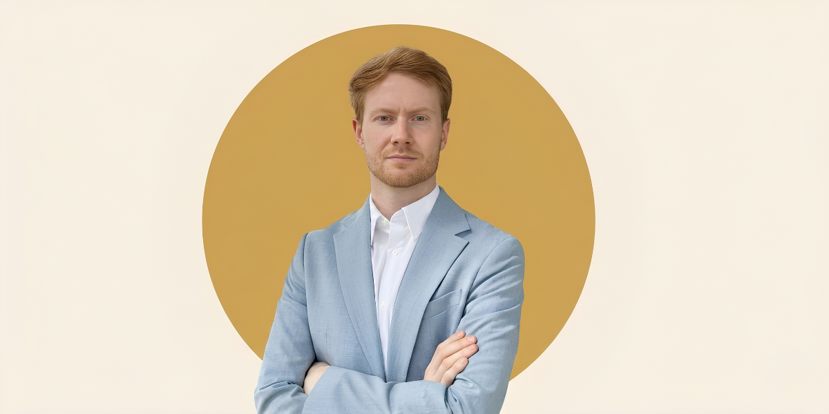 Man in a light blue suit standing with arms crossed against a minimalist gold circular background.